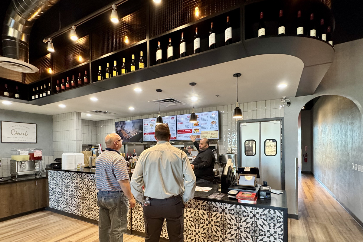 Interior of Pita Kitchen showing customers at the counter ordering food, with menu boards, pendant lighting, and shelves displaying bottles above.