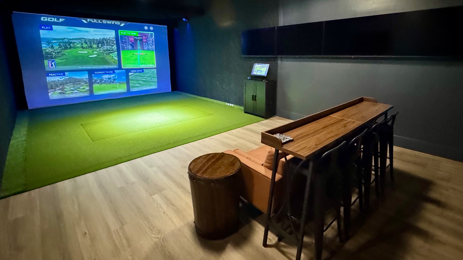 Indoor golf simulator room with a large projection screen, green turf hitting area, and seating with a high table and stools.
