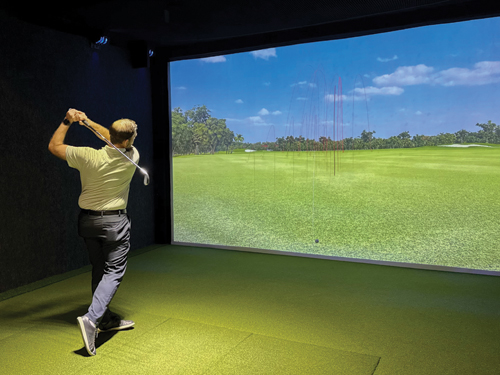 Person swinging a golf club inside a simulator bay with a large screen displaying a virtual golf course.