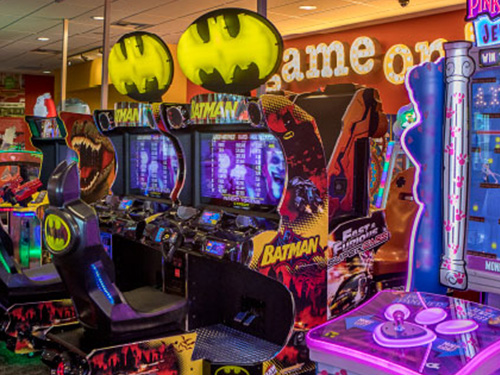 Arcade machines with illuminated bat-symbol signs, surrounded by other colorful games in an arcade.