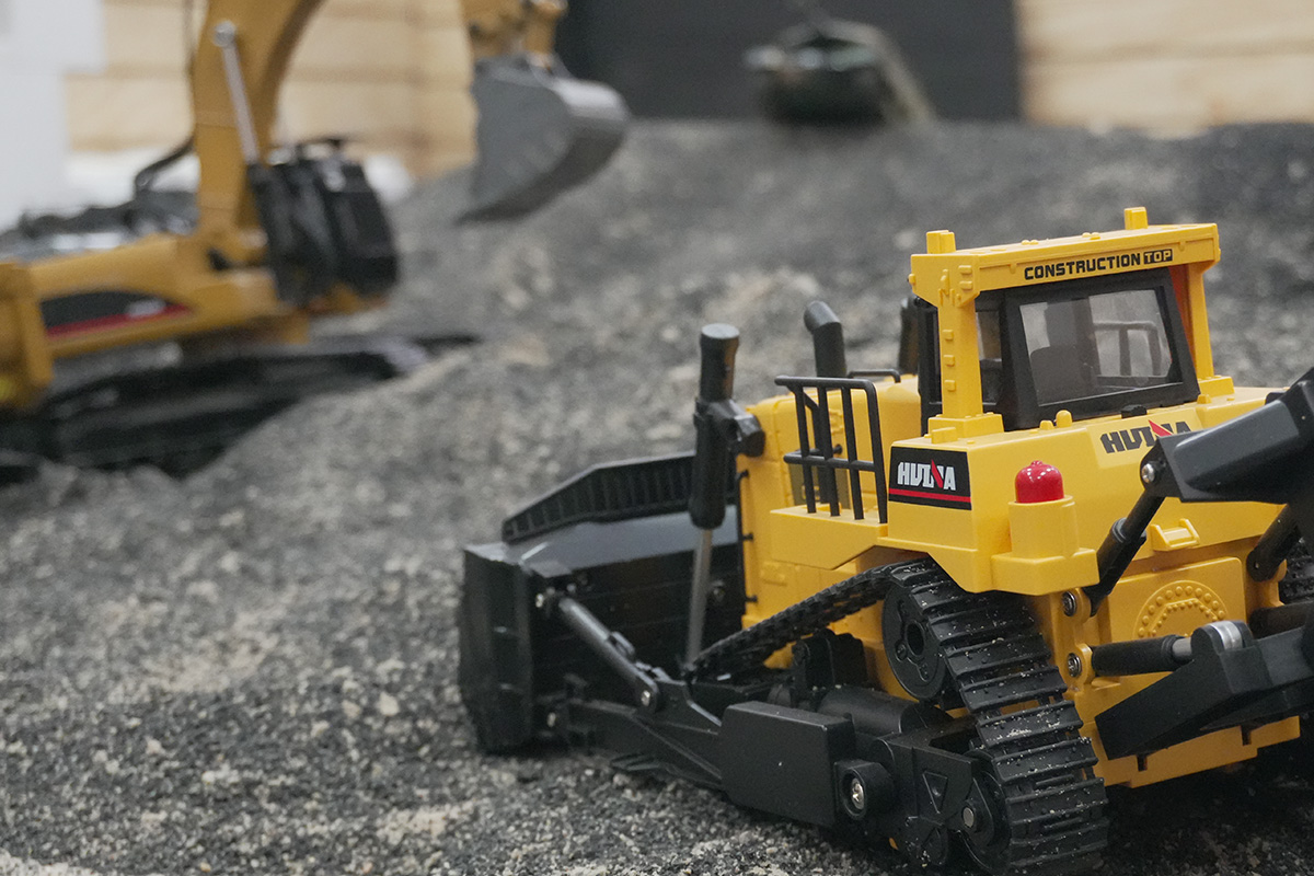 Yellow RC bulldozer with black tracks and a front blade on a gravel-like surface, with an RC excavator blurred in the background.