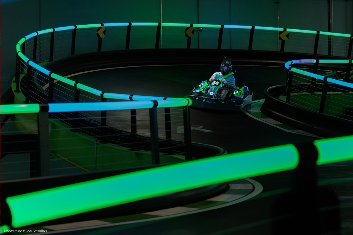 Go-kart driver rounds a curve on an indoor track lined with glowing green and blue barriers.