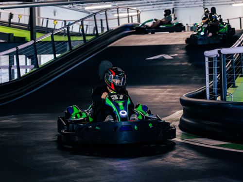 Helmeted go-kart driver races down an indoor track with other karts in the background.