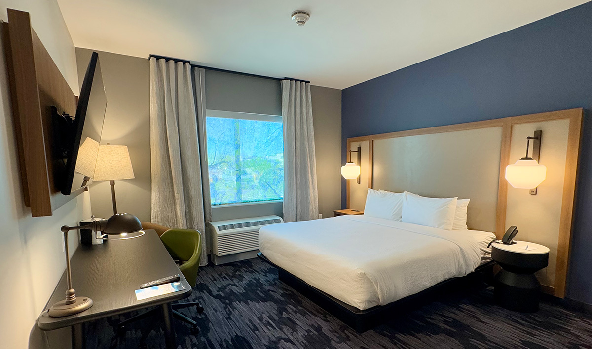 A contemporary hotel room with a king-size bed, blue accent wall, two pendant lights, a work desk with a lamp, and a large window with curtains. The room has a clean, minimalist design with dark carpet flooring.