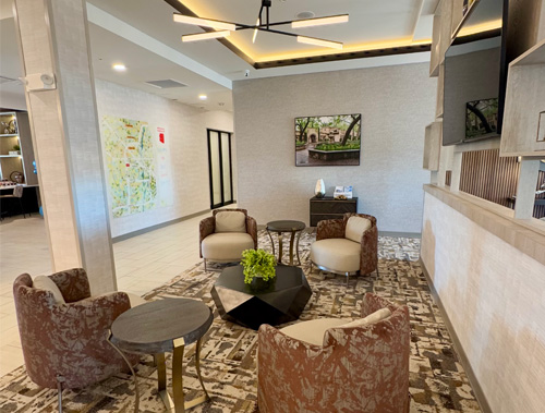 A modern hotel lobby featuring four upholstered armchairs around two small coffee tables, with neutral-toned walls, patterned carpet, and wall art. The space is softly lit and designed for guest relaxation.